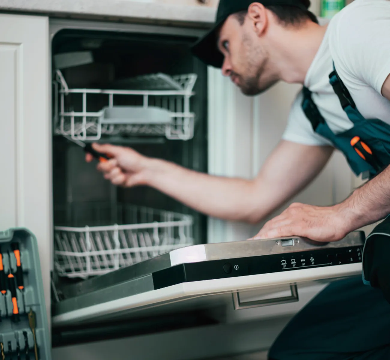 Technician repairing a dishwasher with tools inside a kitchen.
