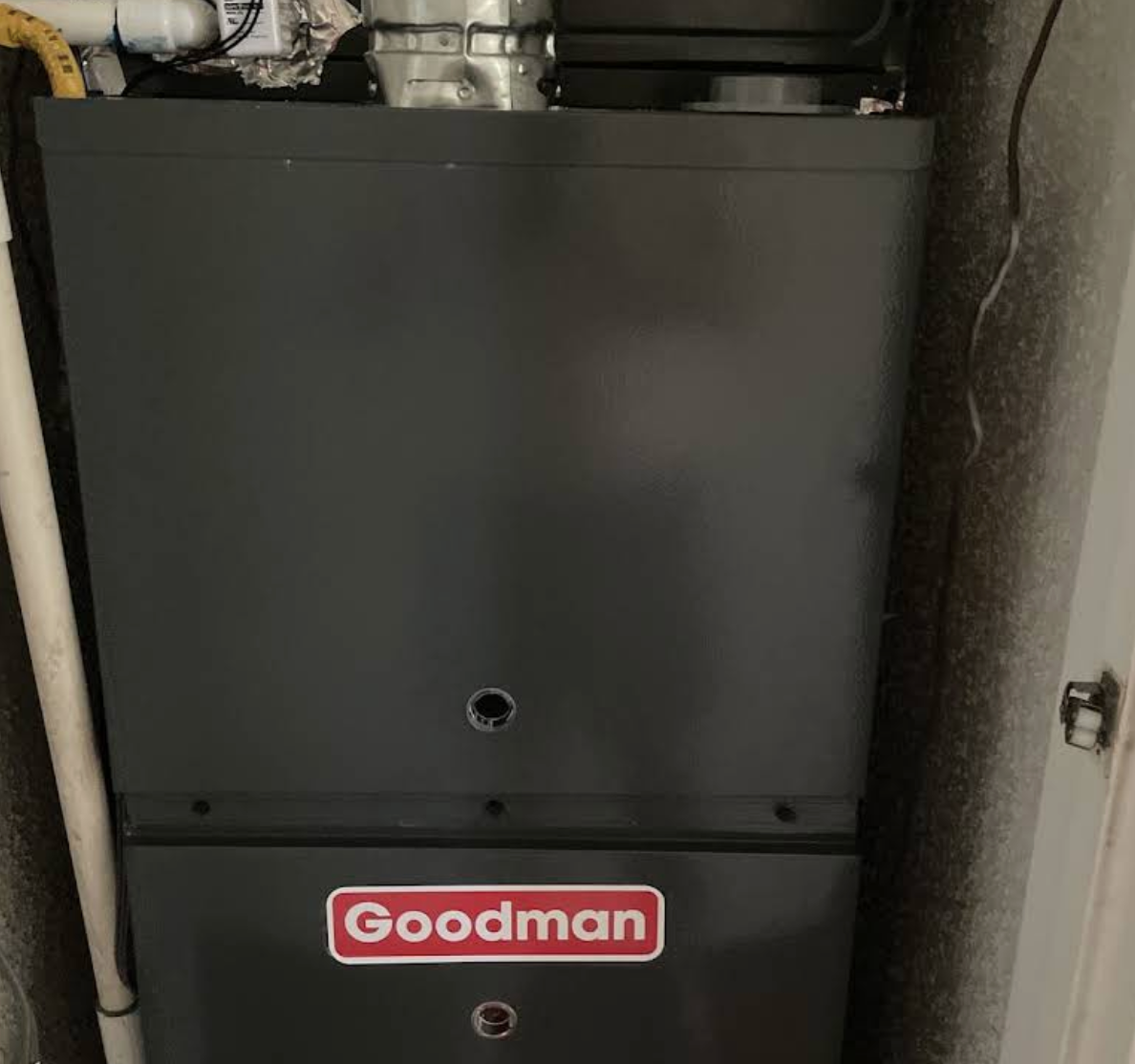 Gray Goodman furnace unit installed between two walls with pipes and cables visible.