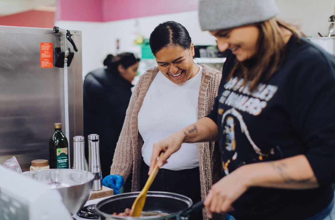 Two women smiling and cooking together in a kitchen with one stirring food in a pan.