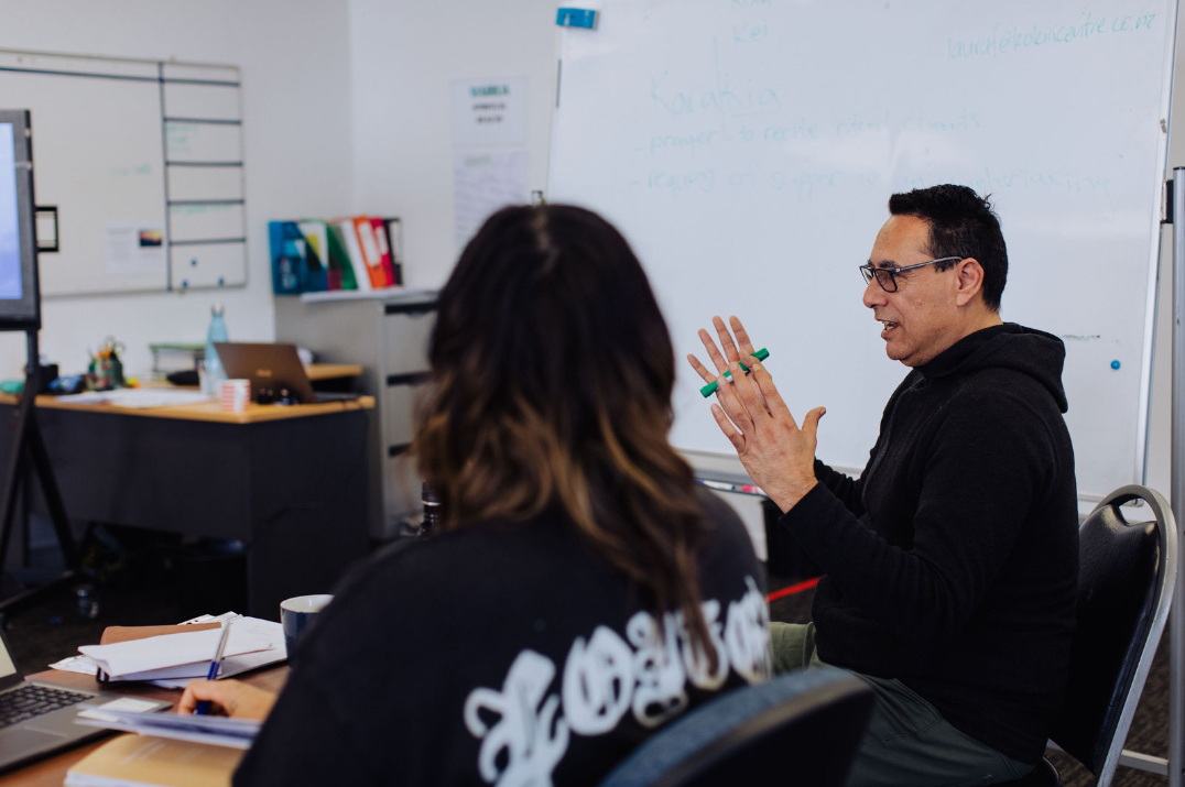 Man in glasses and black hoodie speaking and gesturing with a green marker in a modern office, woman with long hair seen from behind taking notes.