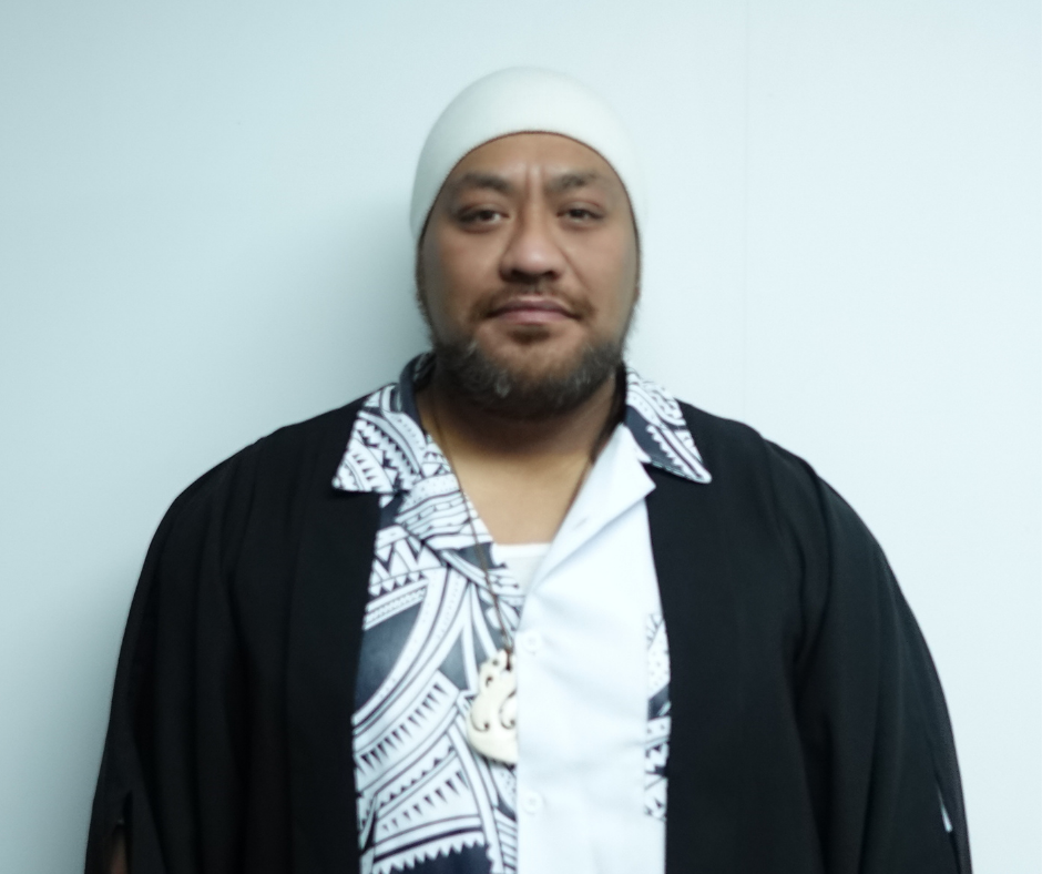 Man wearing a white head covering, patterned black and white shirt, black outer garment, and a carved pendant necklace standing against a plain light background.
