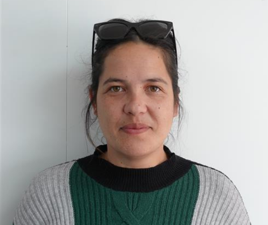 Woman with brown eyes wearing sunglasses on her head and a green and gray sweater standing against a white wall.