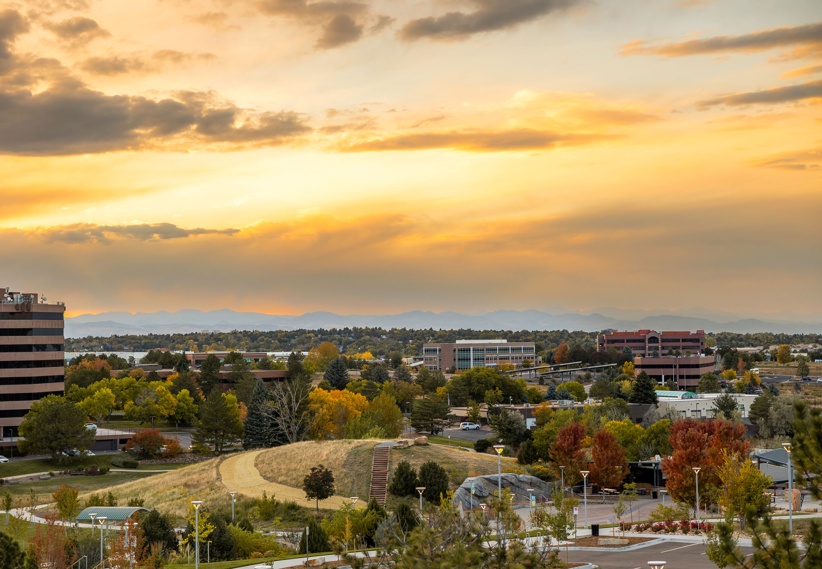 Urban park with autumn trees and buildings under a yellow-orange sunset sky with mountains in the background.
