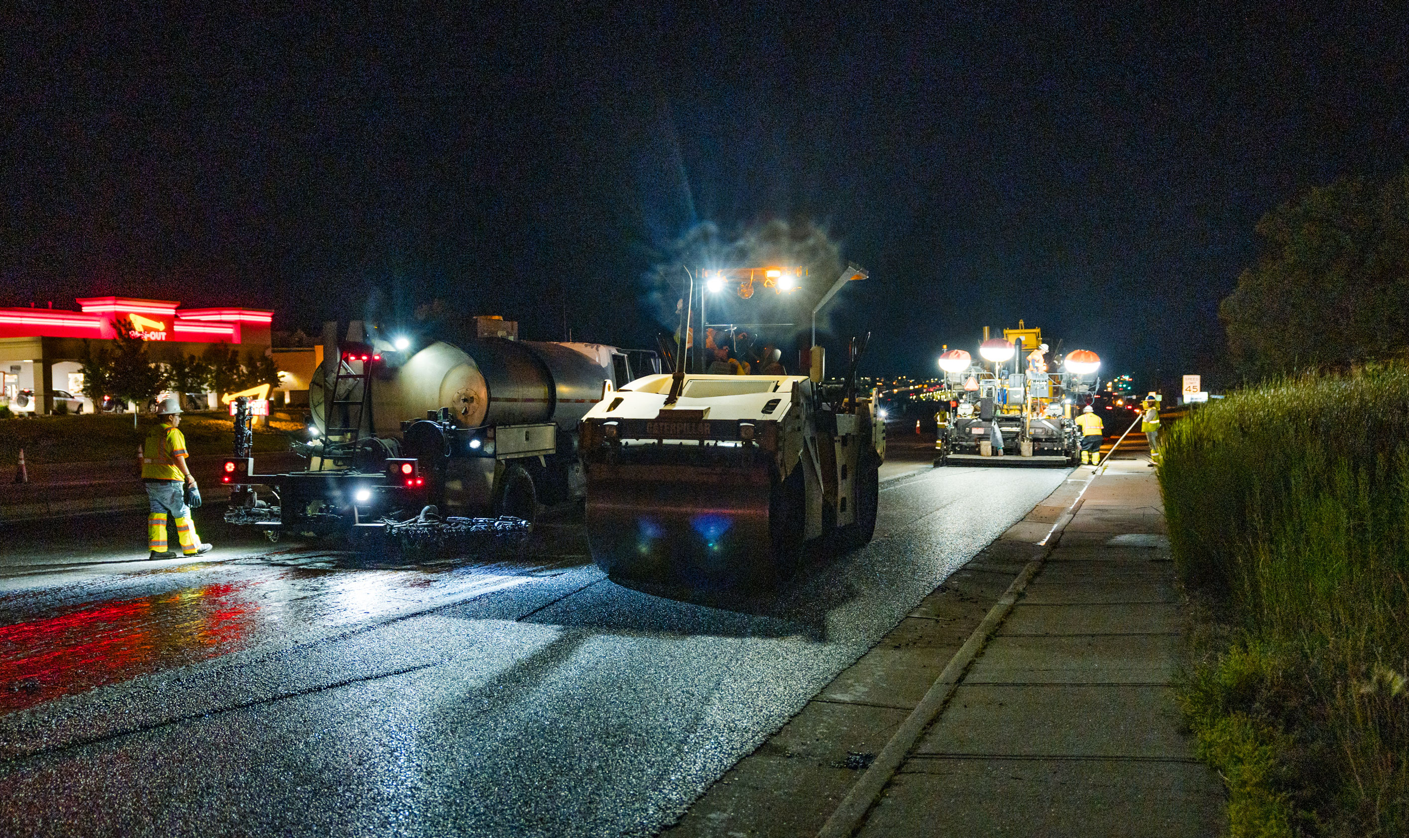 Workers and machinery repaving a road at night with bright lights illuminating the construction area.