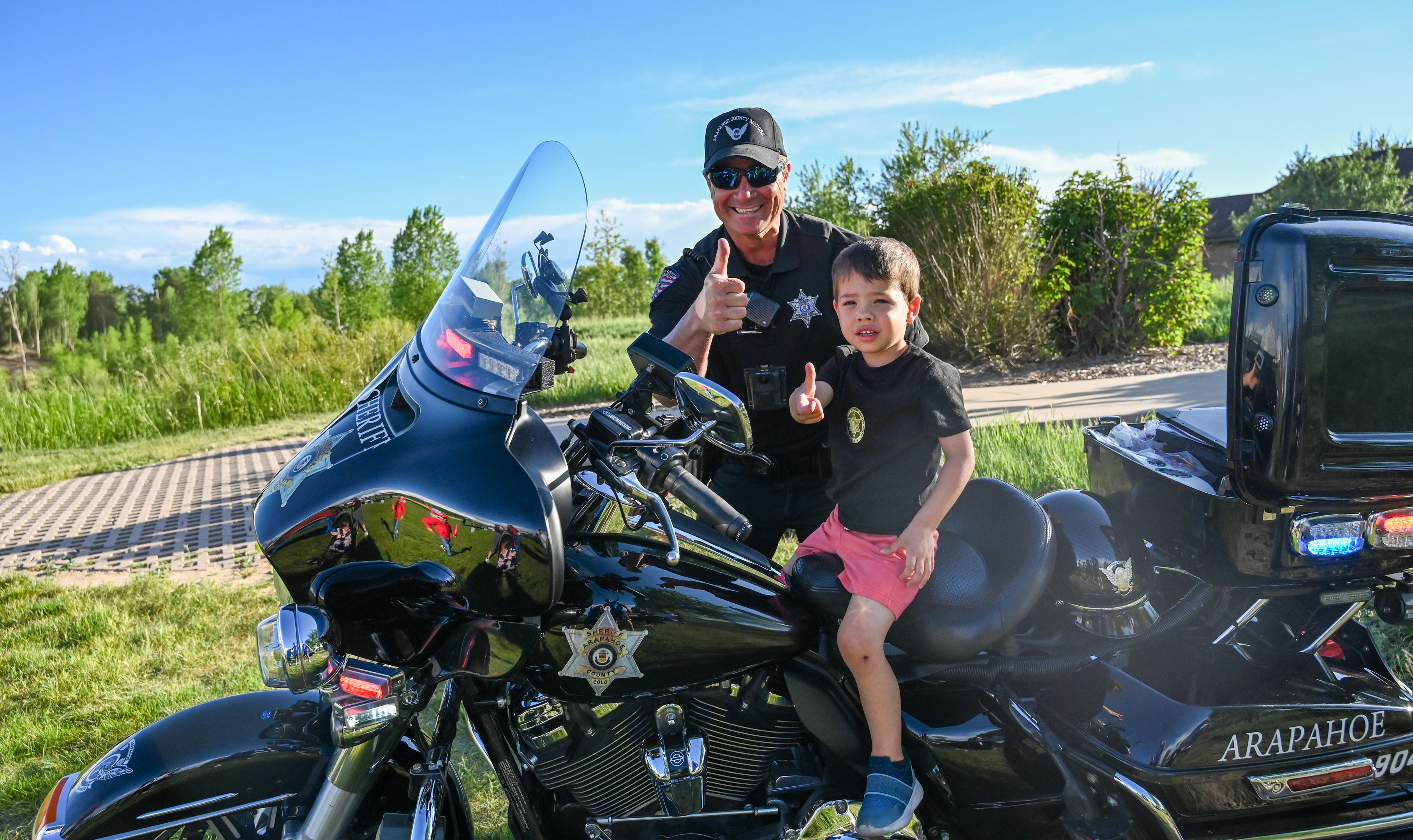 Police officer in sunglasses and cap giving thumbs up next to a young boy sitting on a black Arapahoe sheriff motorcycle, both outdoors on a sunny day.