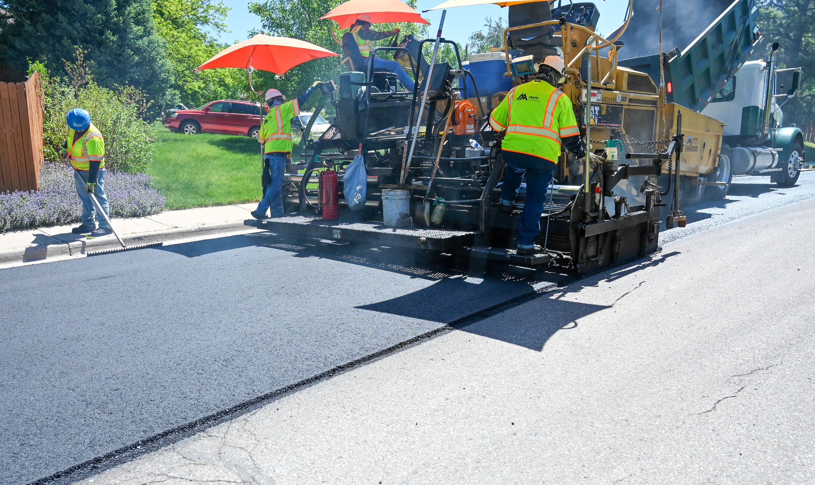 Workers in safety vests repaving a residential street using asphalt paving machinery under orange umbrellas.