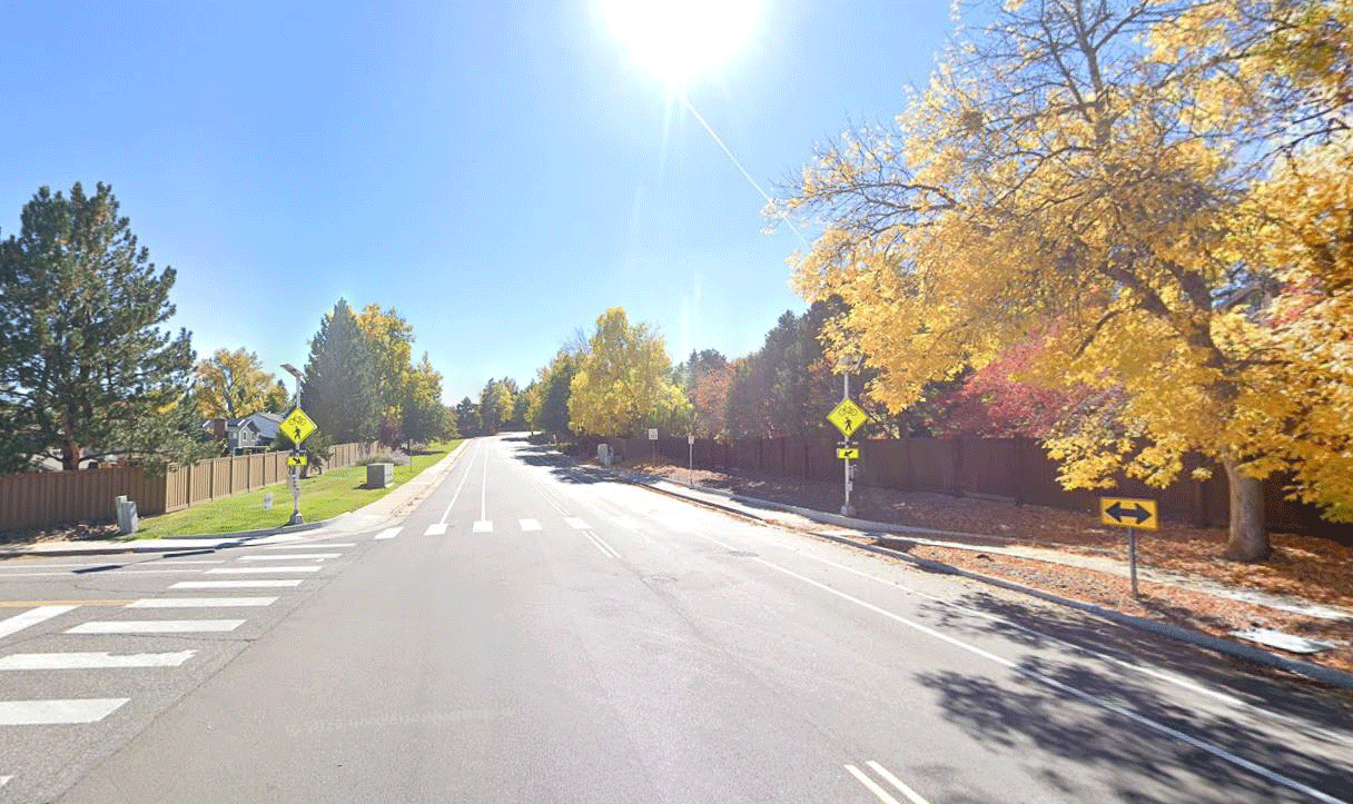 Sunny suburban street with crosswalk, autumn trees, and signs indicating pedestrian crossing and two-way traffic.