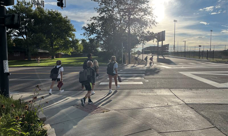 Children with backpacks crossing a street at a crosswalk during late afternoon with sunlight and trees in the background.