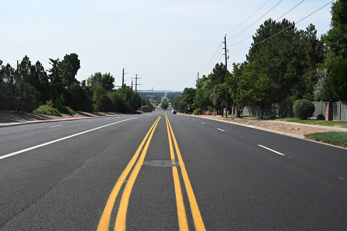 Wide newly paved street with fresh double yellow center lines, flanked by trees and power lines under a clear sky.