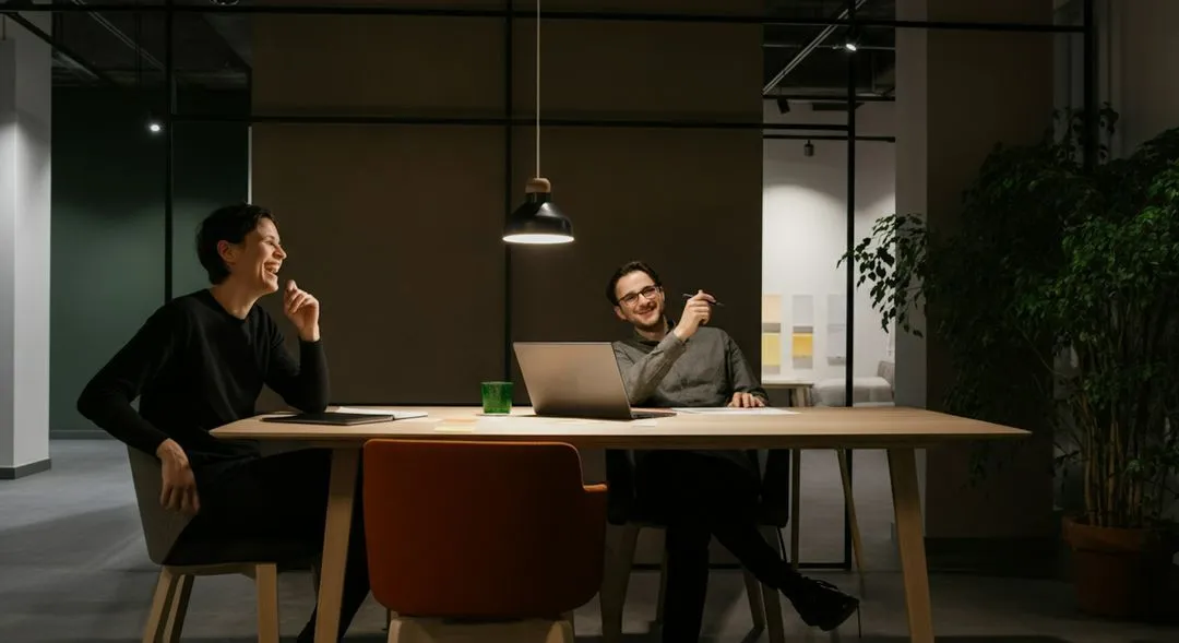Two men sitting at a table with a laptop and a green cup.