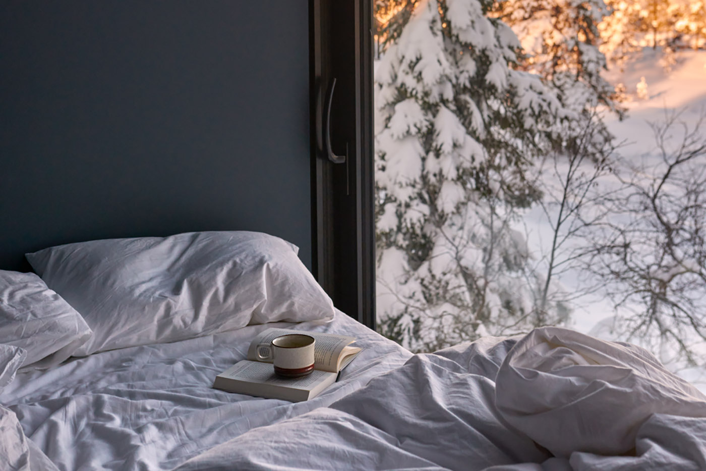 En kopp står på en bok, oppi en seng i en Omgi-hytte med snølandskap utenfor vinduet
