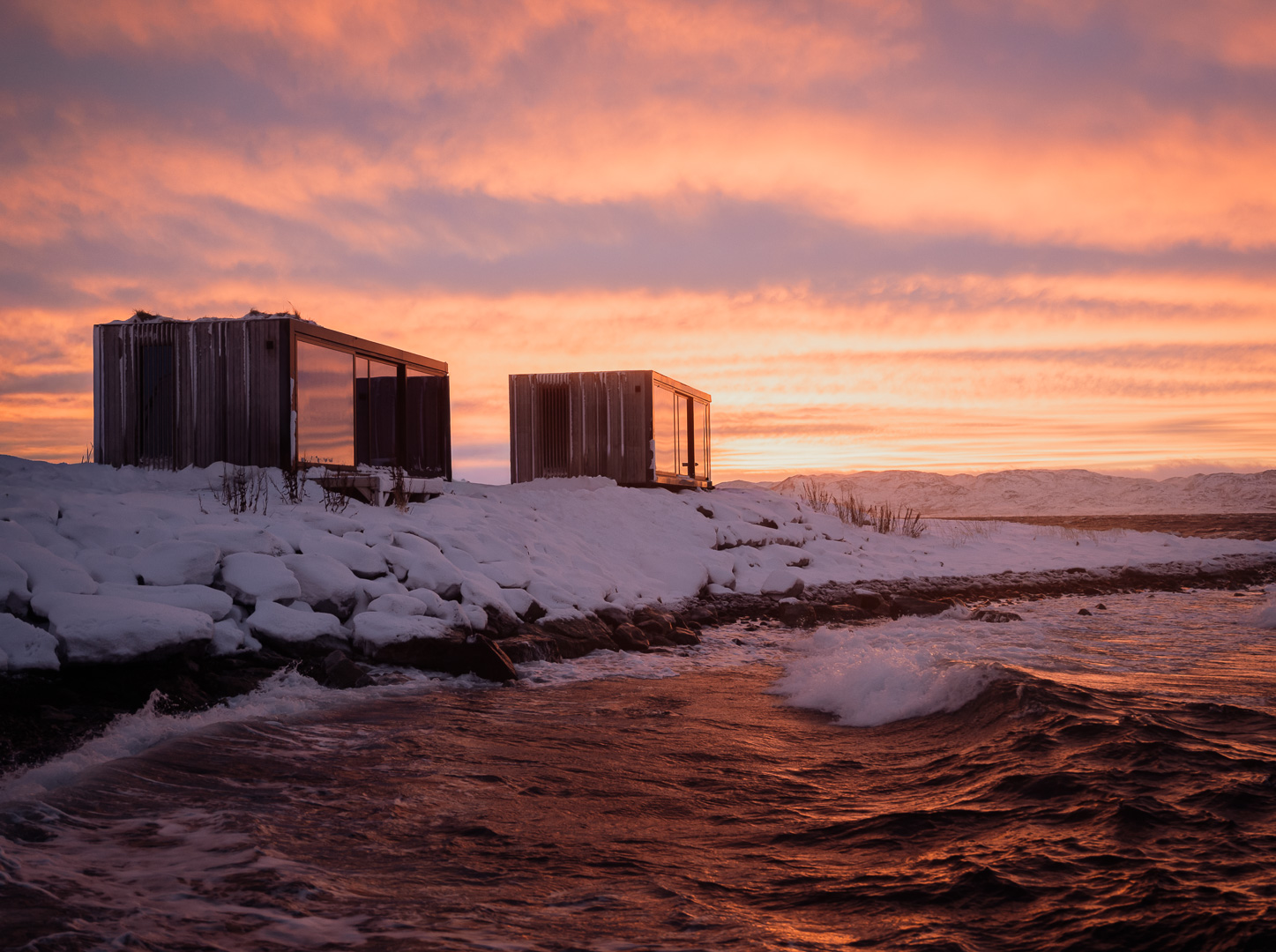 To Omgi hytter i snødekt kystlandskap og rosa himmel