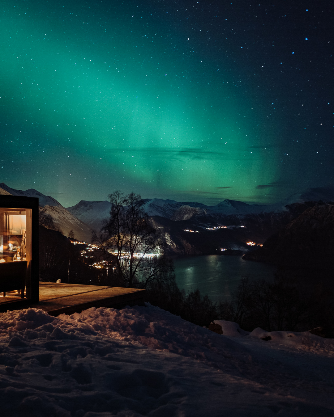 Nordlys på himmel over fjell og fjord, med en hytte i forgrunnen
