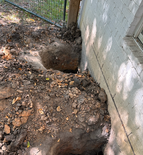 A deep excavation is visible next to a white brick building wall and a chain-link fence. — Texas Greatest Remodelers