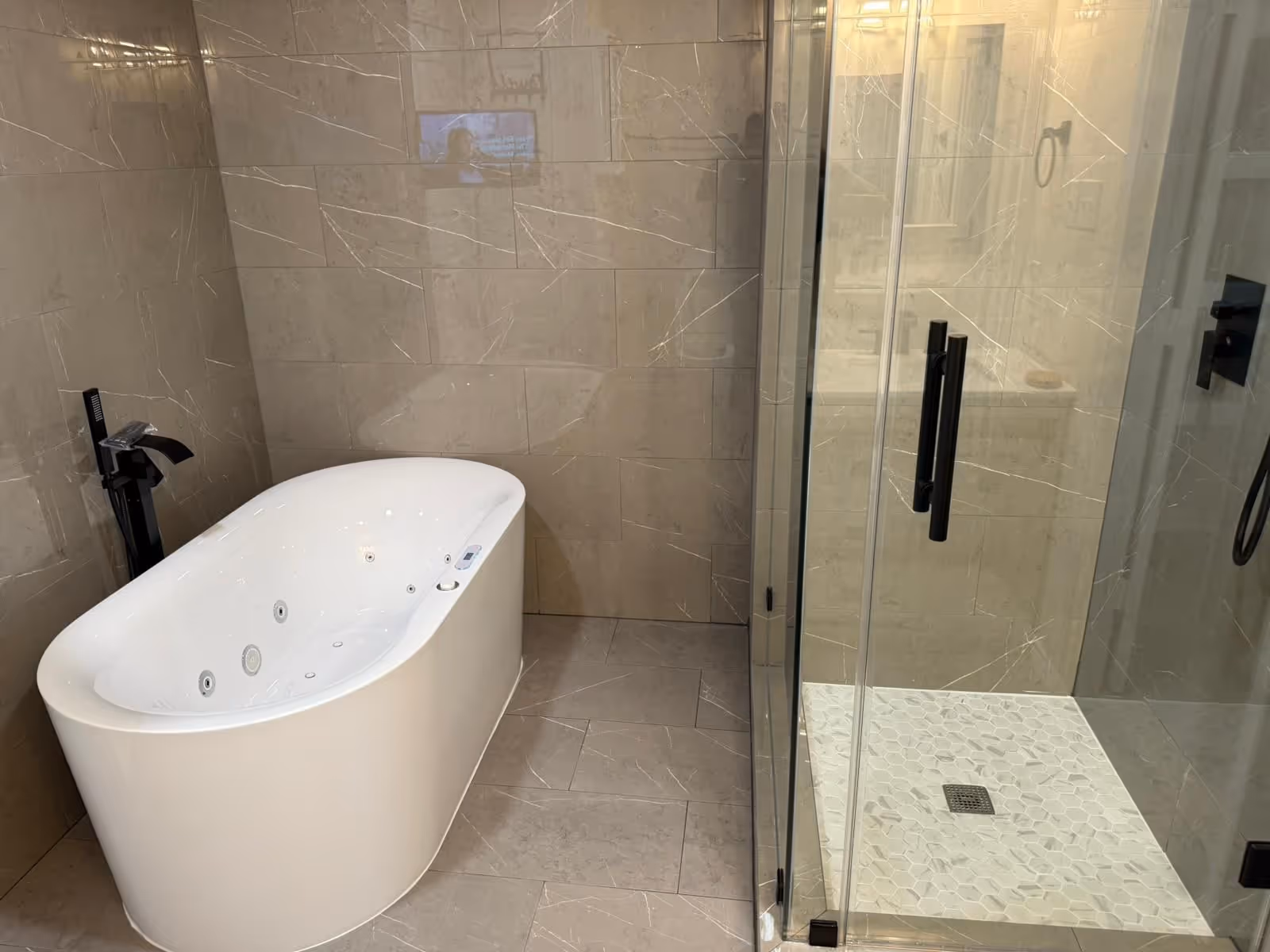A modern bathroom features a white freestanding jetted soaking tub set against beige marble-patterned wall tiles next to a gl