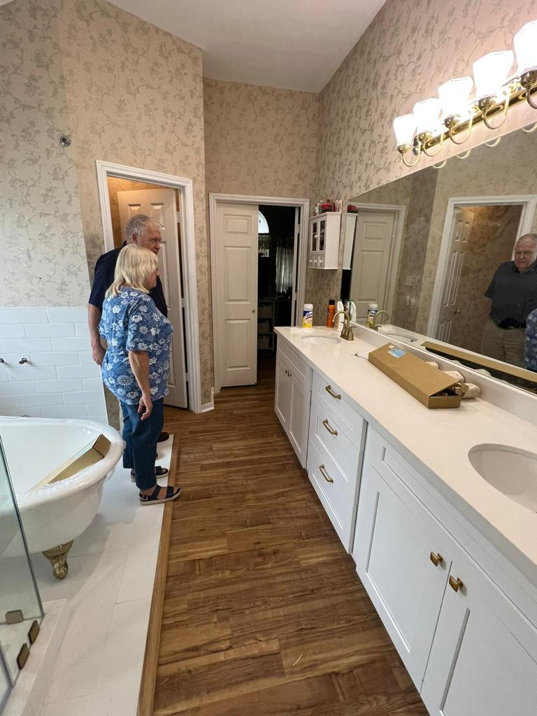Two people are inspecting a renovated bathroom featuring white cabinetry, a large mirror, and wood-look flooring. — Texas Gre
