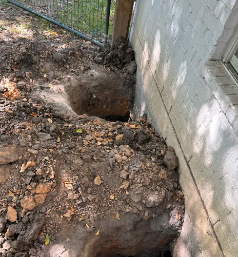 A deep excavation is visible next to a white brick building wall and a chain-link fence. — Texas Greatest Remodelers