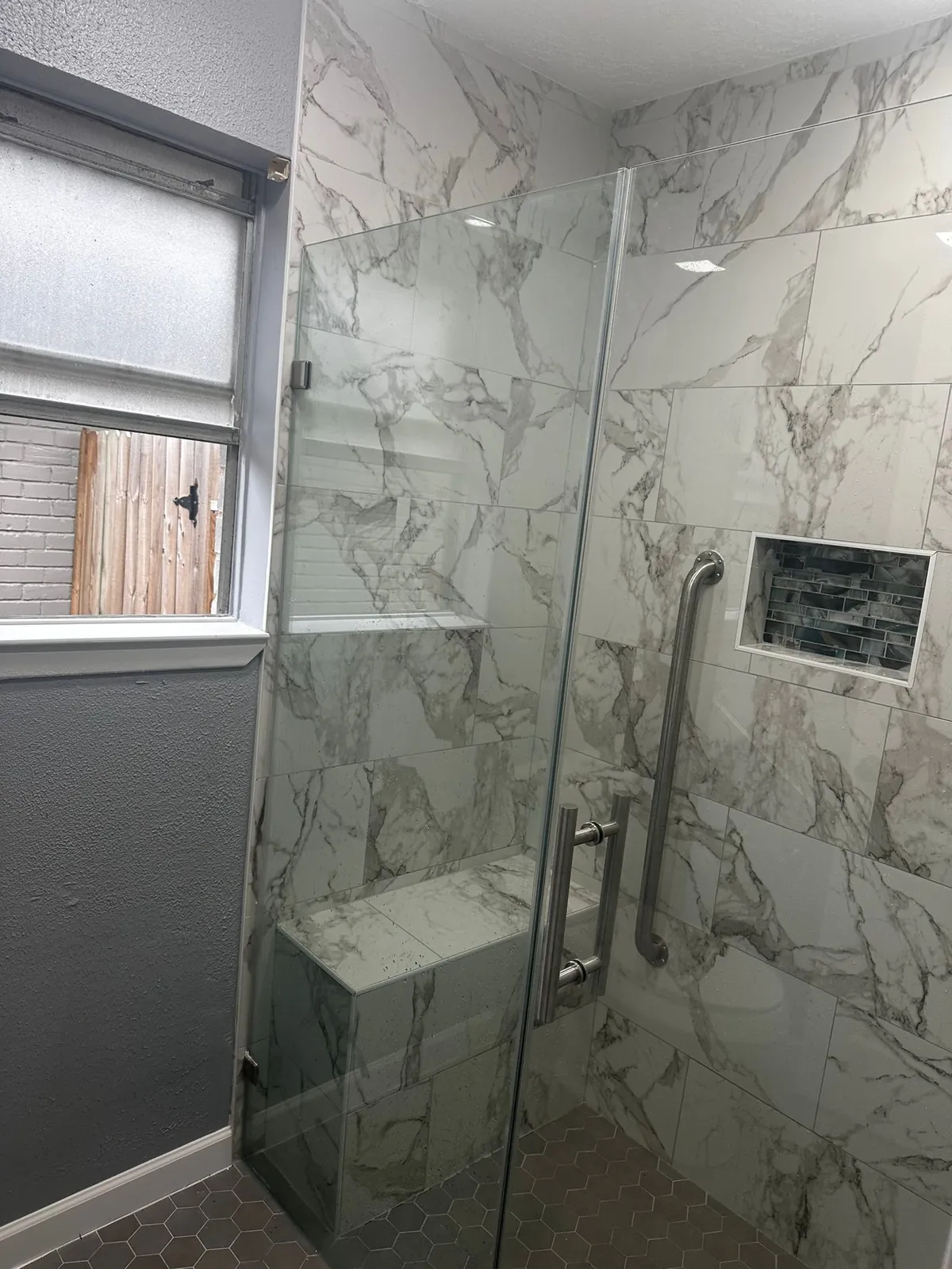 A modern walk-in shower features large marble-patterned wall tiles, a built-in marble bench, a glass enclosure with brushed n