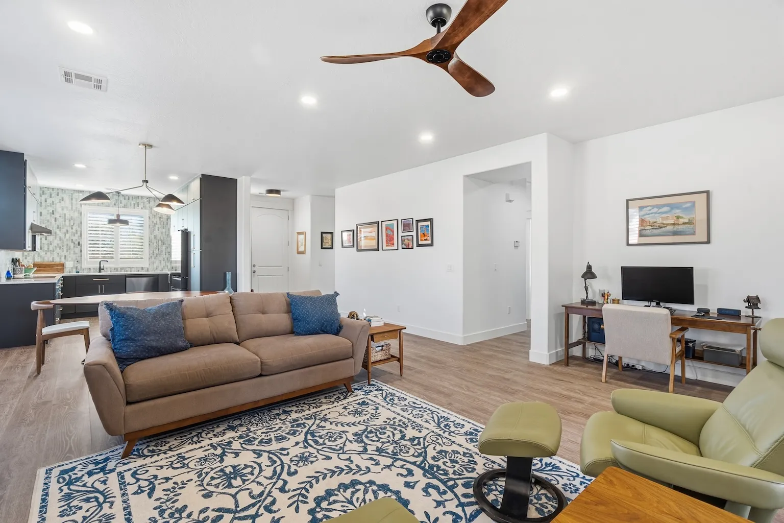 Modern open-concept living area with a beige sofa, blue patterned rug, green armchair with footrest, and an adjacent kitchen with black cabinetry and mosaic backsplash.