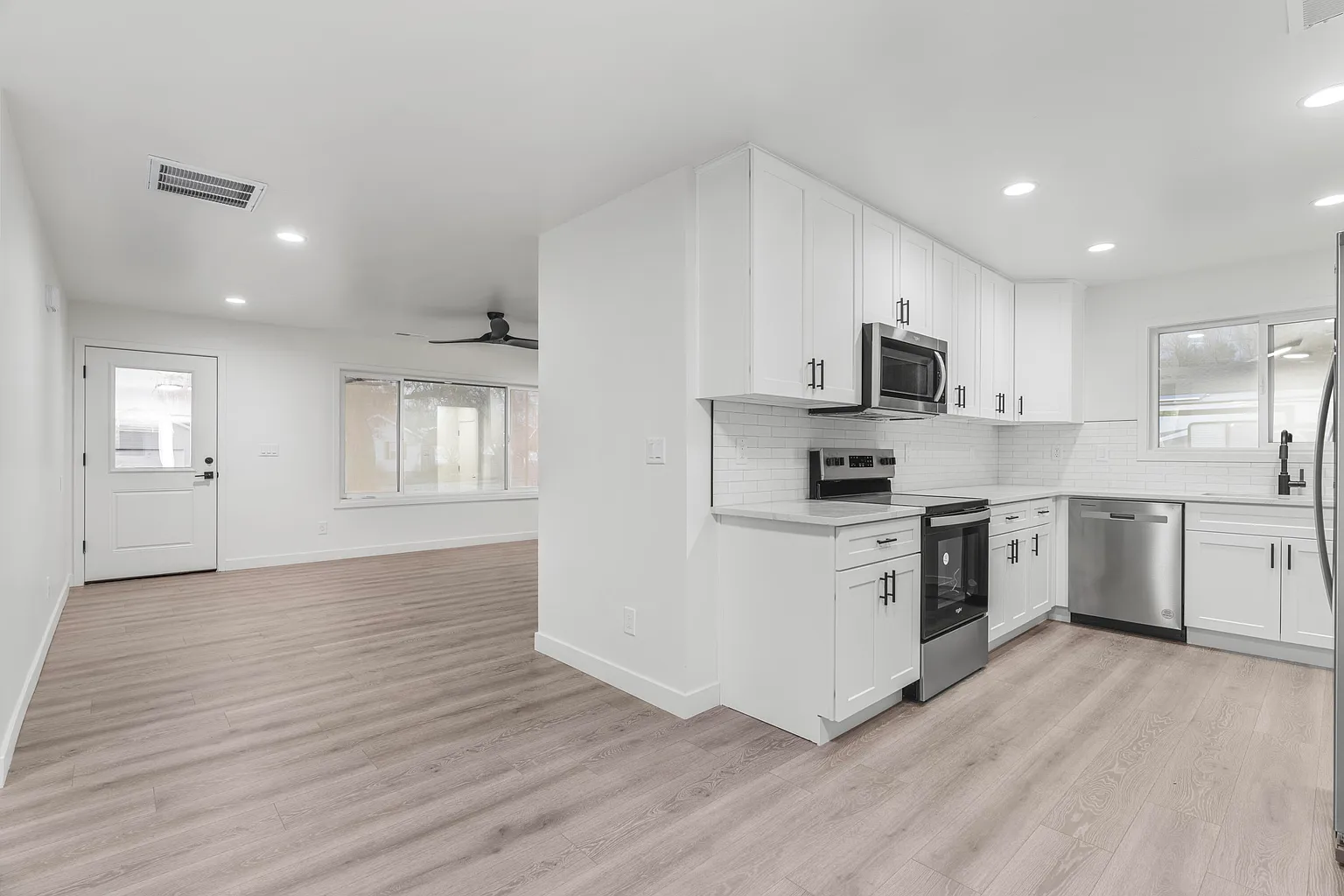Modern kitchen with white cabinets, stainless steel appliances, and light wood flooring adjacent to an empty living area with a large window and white door.