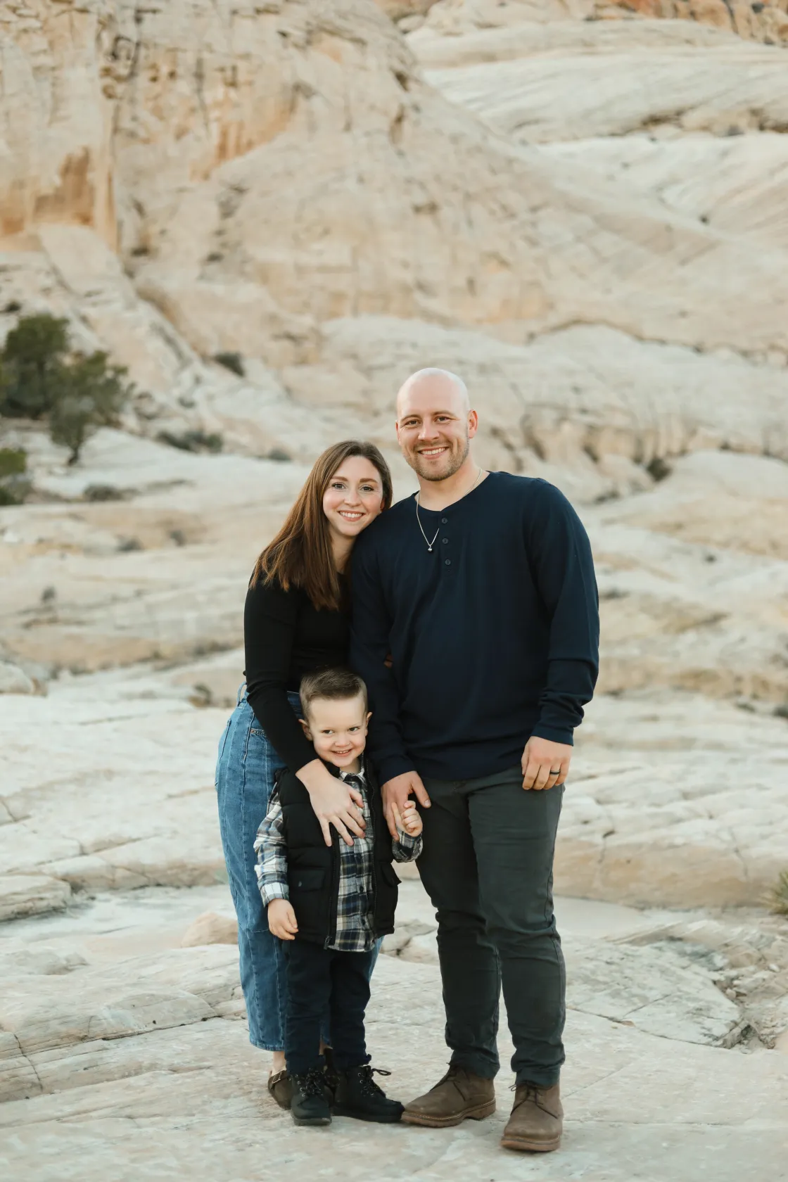 Smiling family of three standing on rocky terrain with a mountain backdrop, including a man, woman, and young boy.