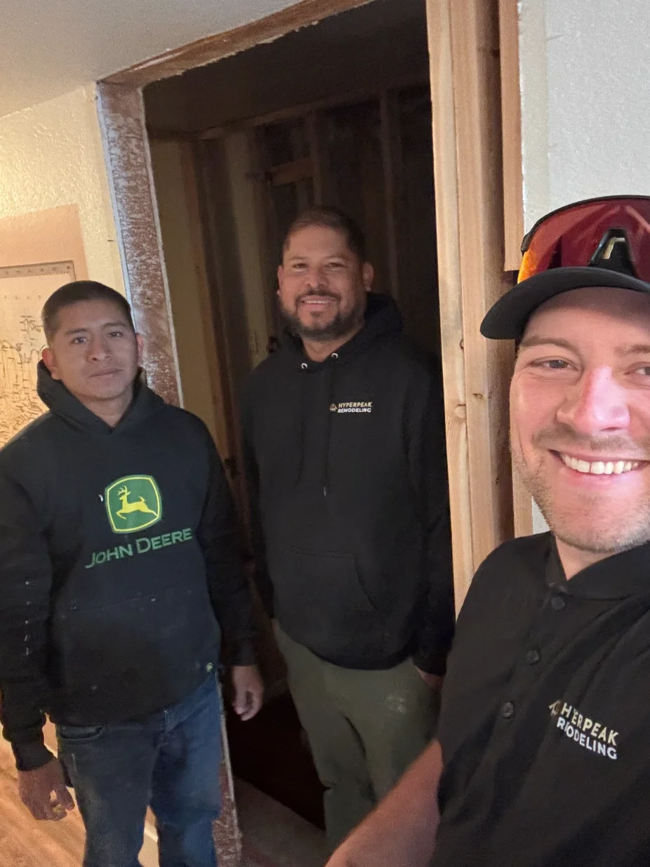Three men smiling indoors near a doorway, two wearing Hyperpeak Remodeling hoodies and one in a John Deere hoodie.