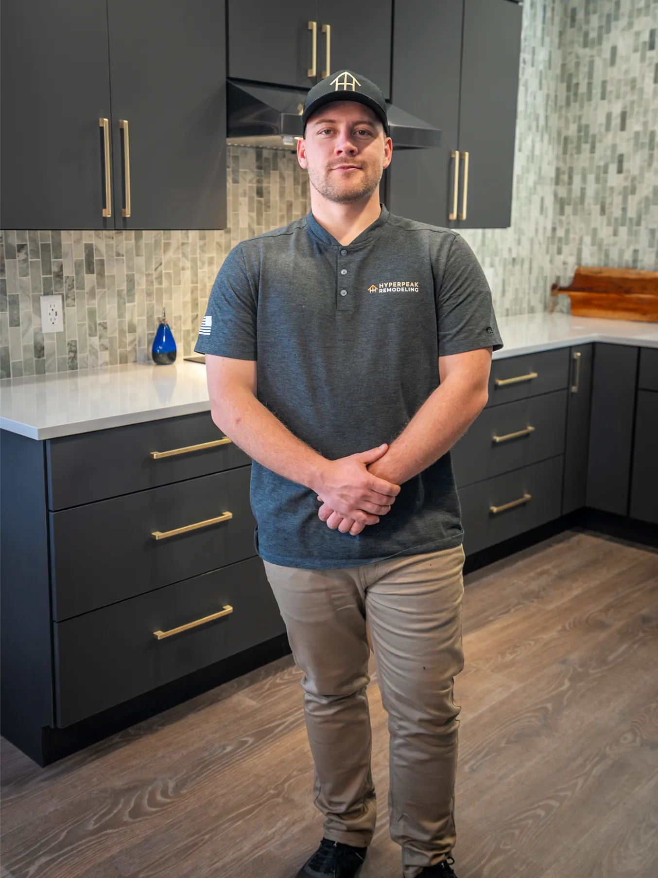 Man wearing a dark gray Hyperpeak Remodeling shirt and hat stands in a modern kitchen with gray cabinets and a mosaic tile backsplash.