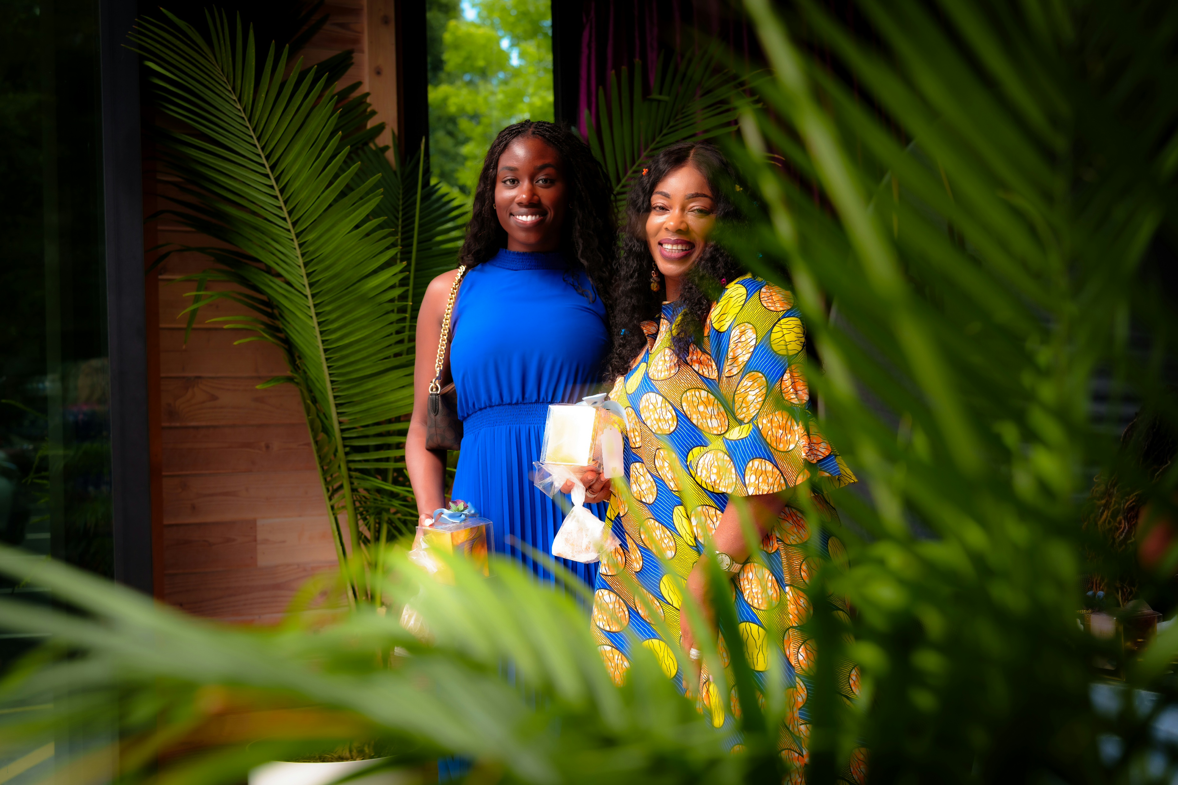 Bridal shower portrait of two smiling women standing among green palm leaves. One wears a blue dress, and the other wears a colorful patterned dress while holding gifts. Captured by HAKIM Studios, highlighting joyful and candid moments.