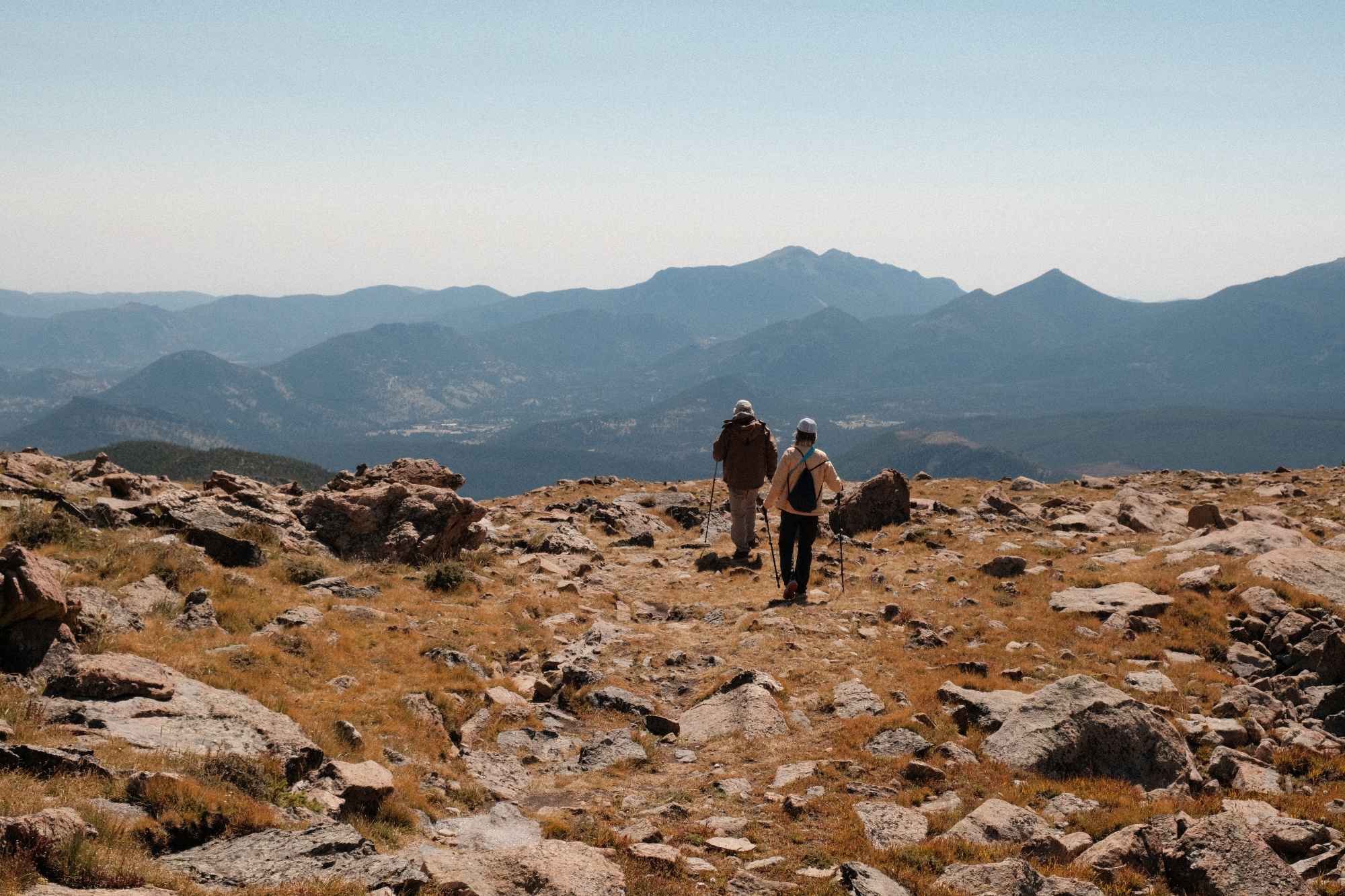Two hikers walking with trekking poles on a rocky mountain trail with distant mountain ranges under a clear sky.