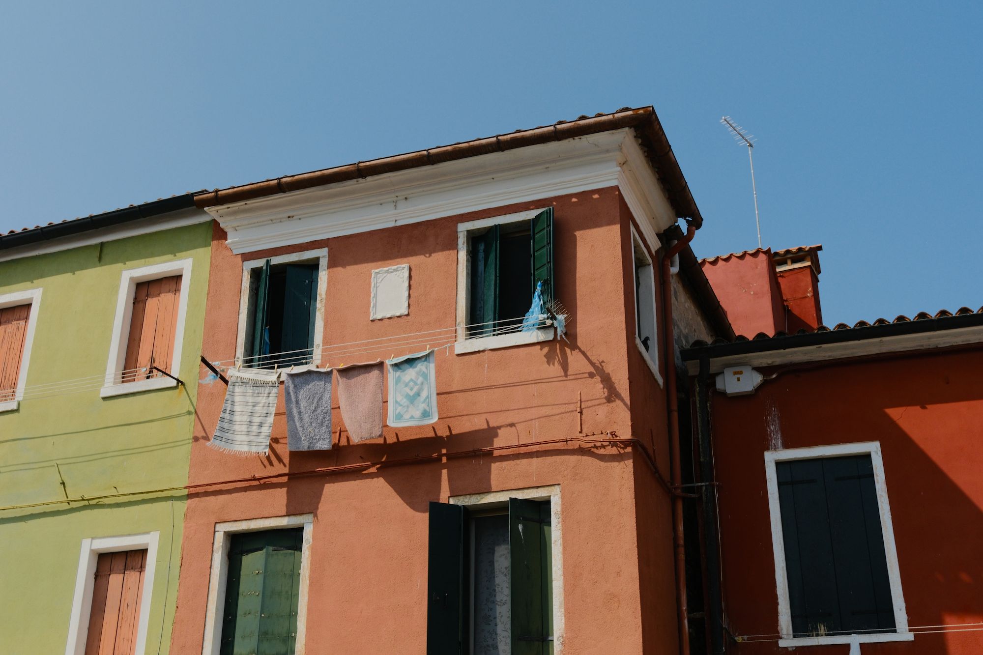 Colorful Italian-style buildings with green shutters and laundry hanging on a clothesline under a clear blue sky.