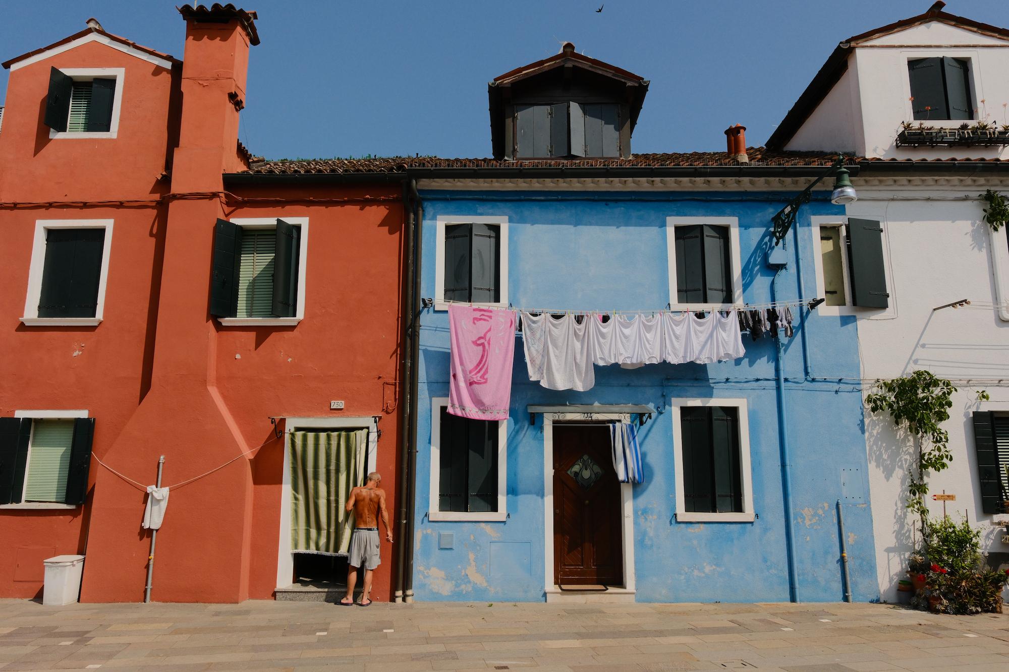 Colorful row of houses with red, blue, and white facades, laundry hanging between windows, and a shirtless man standing near a green-striped curtain door.