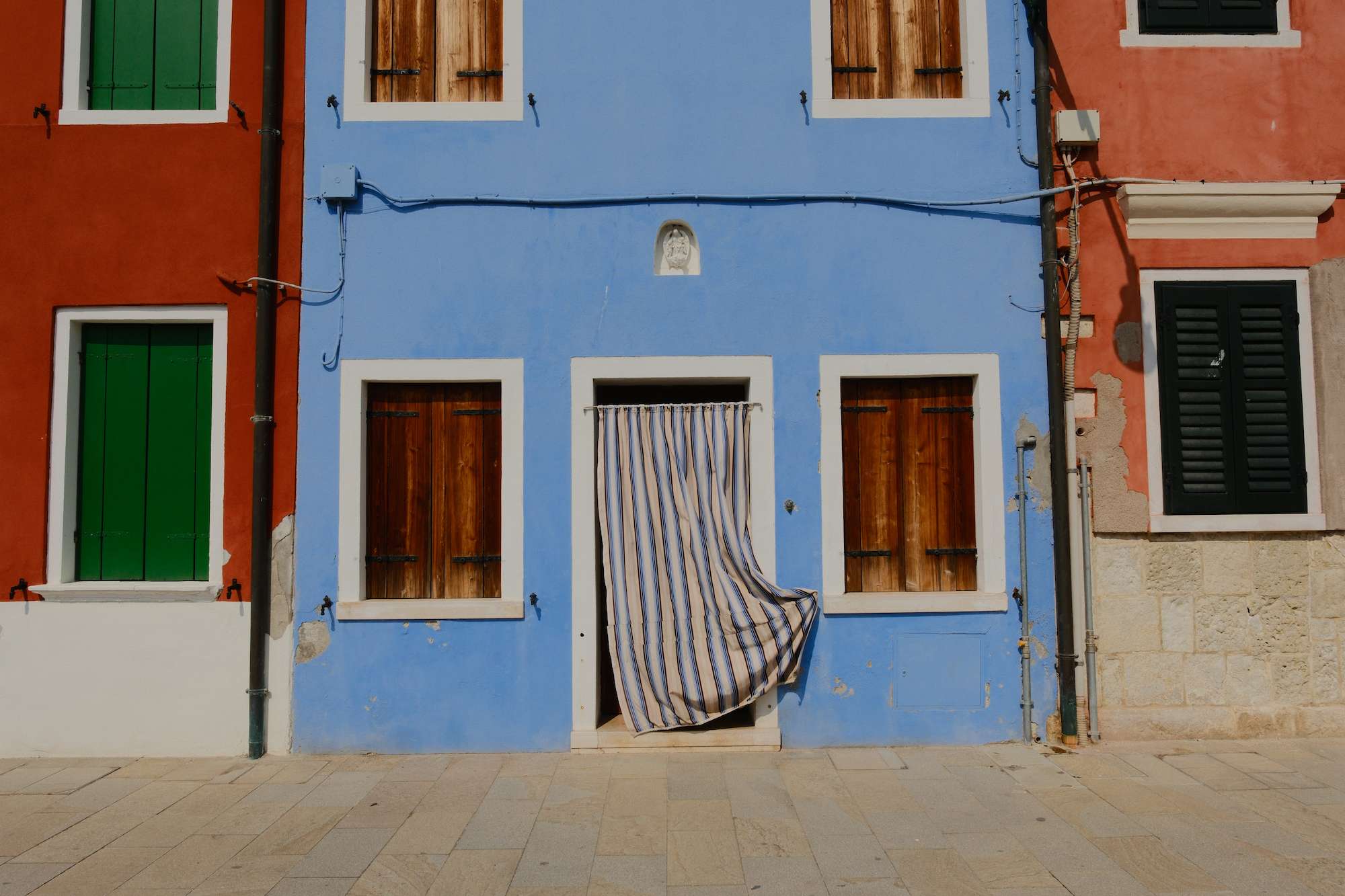 Colorful row of buildings with blue facade in the center featuring wooden shuttered windows and a doorway covered by a flowing striped curtain.
