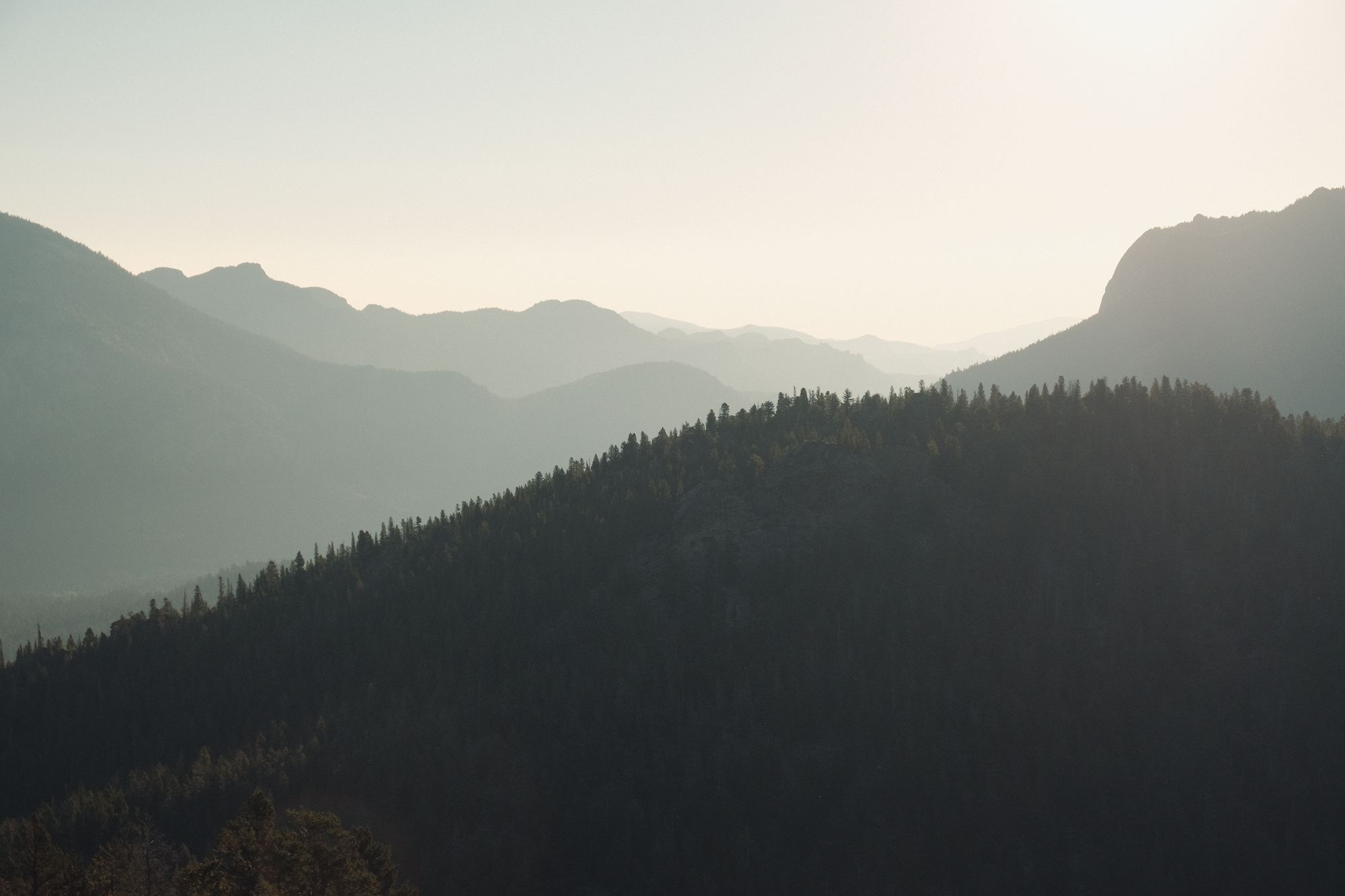 Layered mountain ranges with a forested hill in the foreground under a hazy sky.