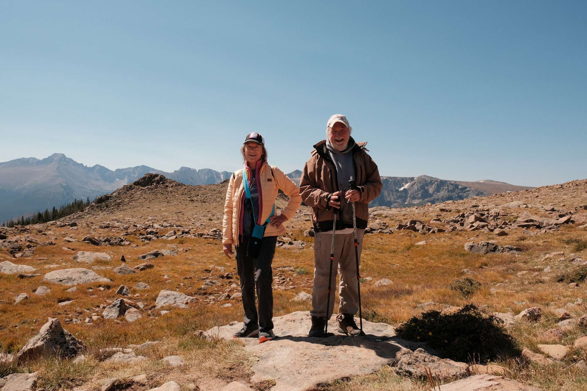 Two smiling elderly hikers standing on a rocky mountain meadow with hiking poles under a clear blue sky.