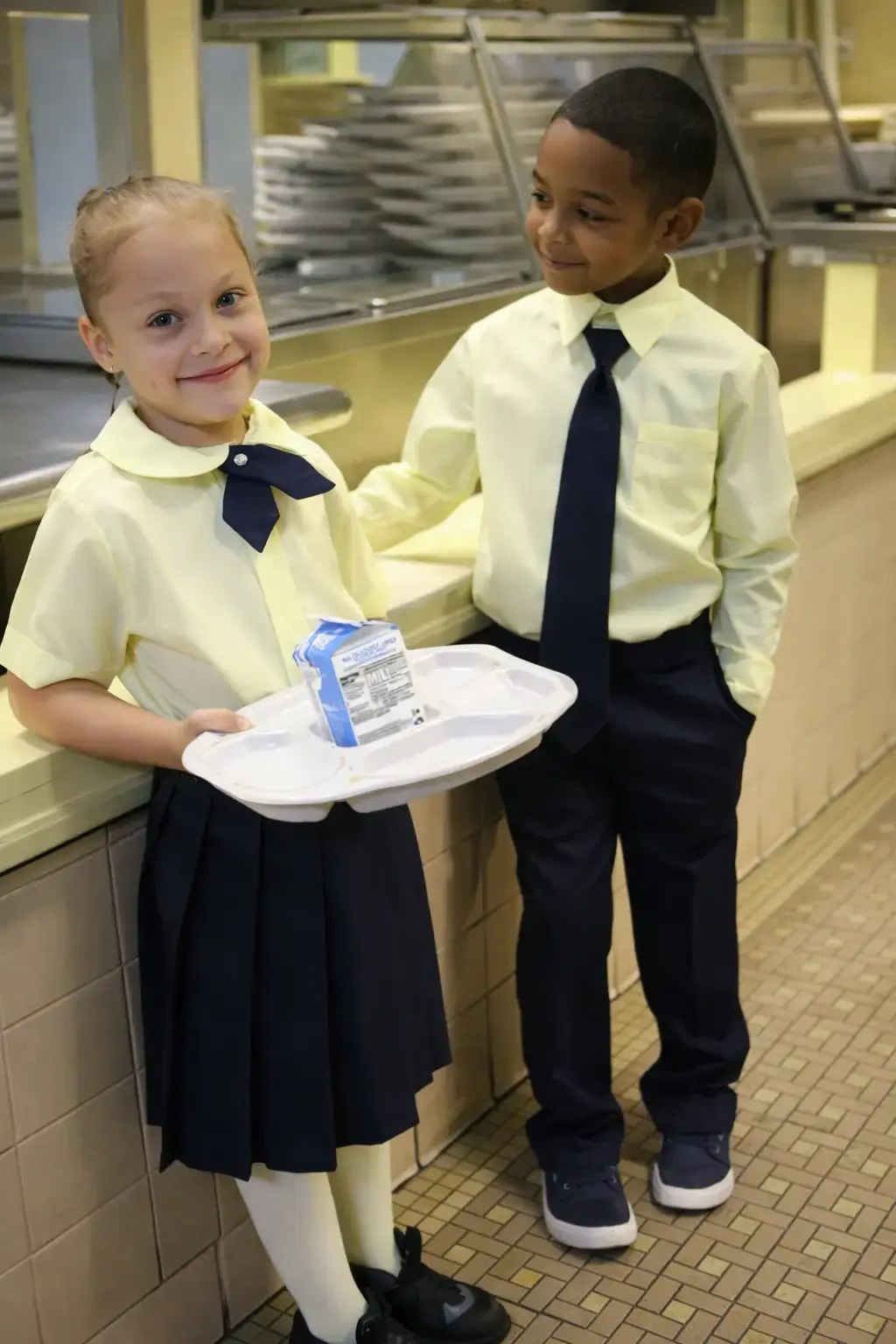 Two students in school uniforms holding lunch tray in cafeteria