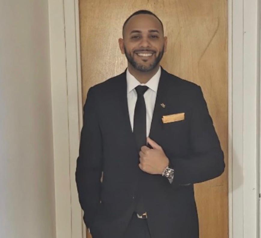 Man smiling wearing black suit, white shirt, black tie, orange pocket square, and wristwatch.