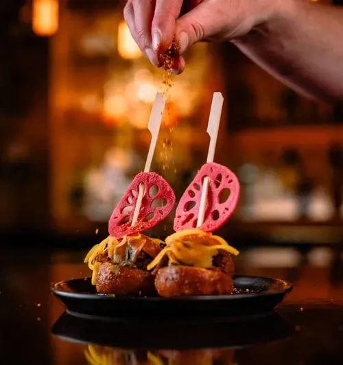 Two gourmet appetizers with thin pink chips on wooden sticks topped with shredded cheese, being sprinkled with seasoning by a hand.