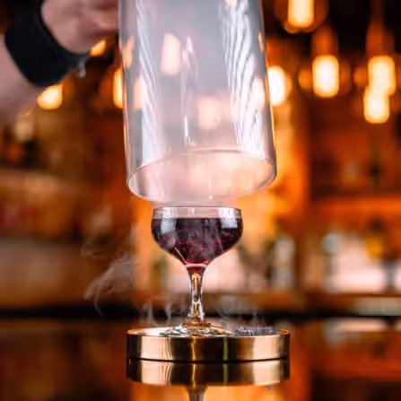 A hand lifting a glass dome from a stemmed glass of red wine creating a smoky effect on a wooden surface with warm bokeh lights in the background.