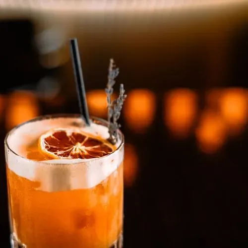 Orange cocktail in a glass with a black straw, garnished with a dried citrus slice and a sprig of herbs.