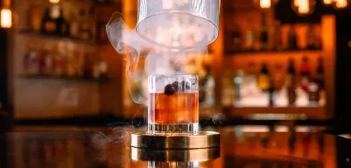 Whiskey glass with smoke rising inside a cylindrical smoke infuser on a bar counter with blurred bottles in background.