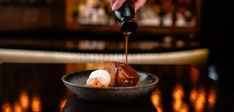 Chocolate cake slice on a plate with cream and honeycomb, chocolate sauce being poured over it.
