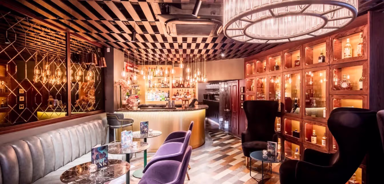 Elegant bar interior with leather banquette, purple chairs, illuminated liquor shelves, and hanging pendant lights.
