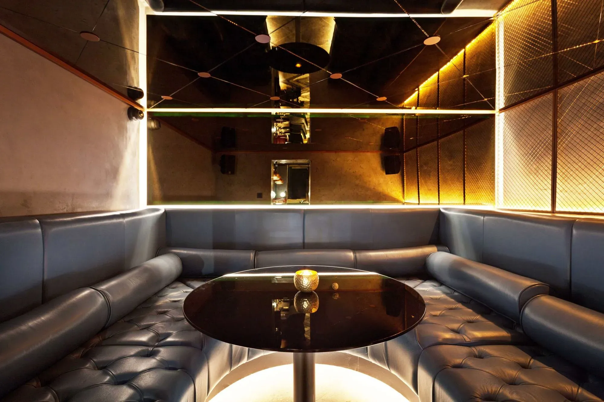 Cozy lounge area with tufted black leather U-shaped seating around a circular black table with a small golden candle holder on top.