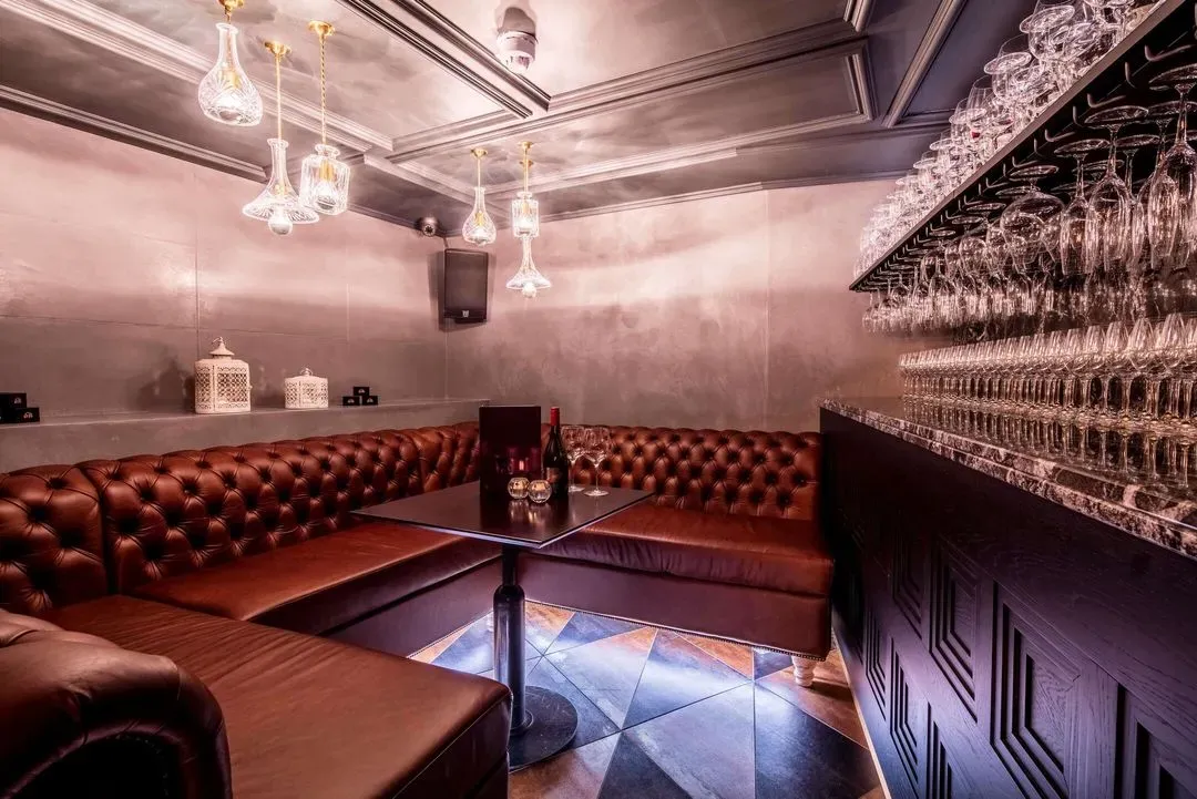 Cozy corner seating area with brown tufted leather couches around a small table holding a wine bottle and glasses, next to a bar with hanging wine glasses.