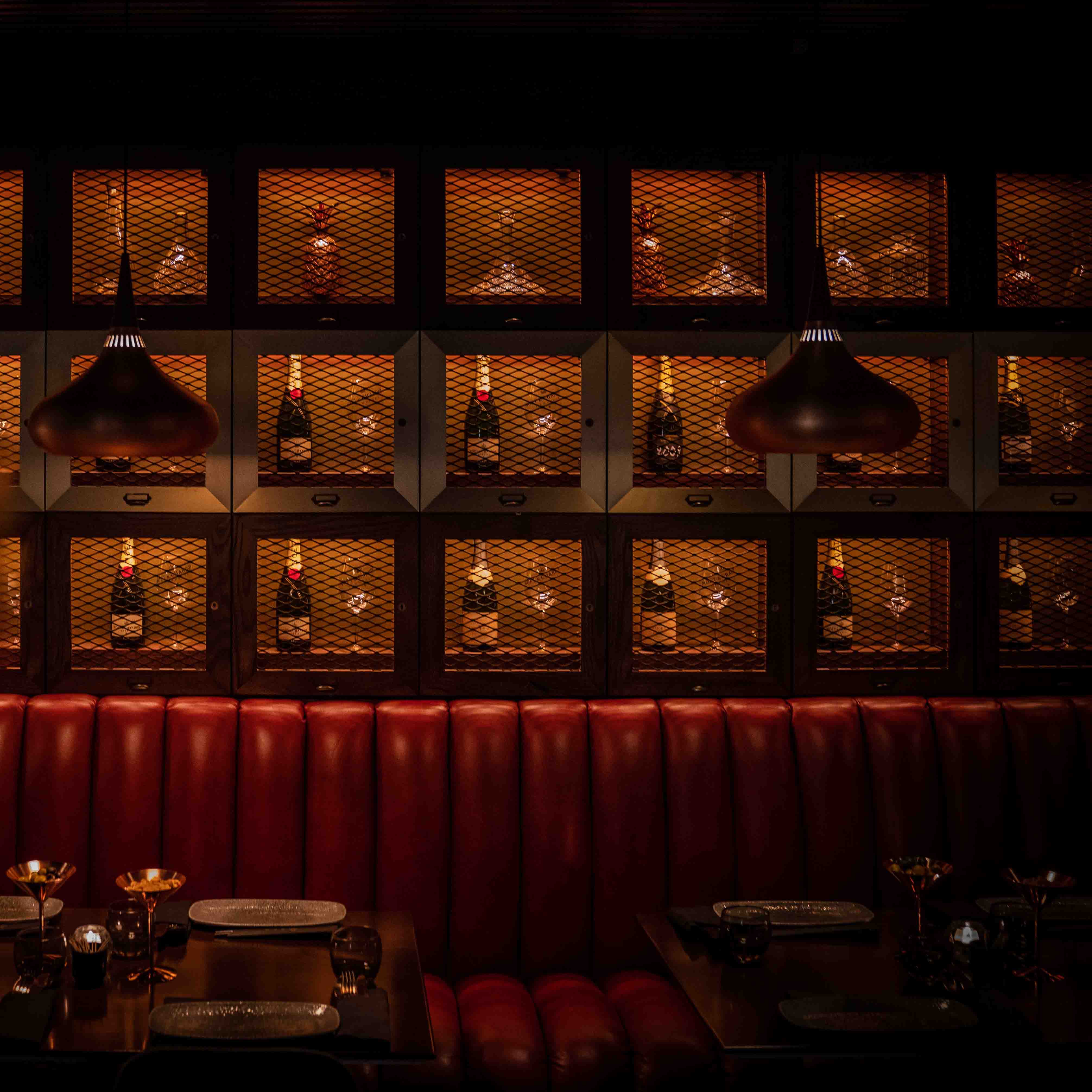 Dimly lit restaurant booth with red leather seating, tables set with gold-rimmed glasses and plates, back wall featuring illuminated shelves with bottles and decorative glassware behind mesh panels.