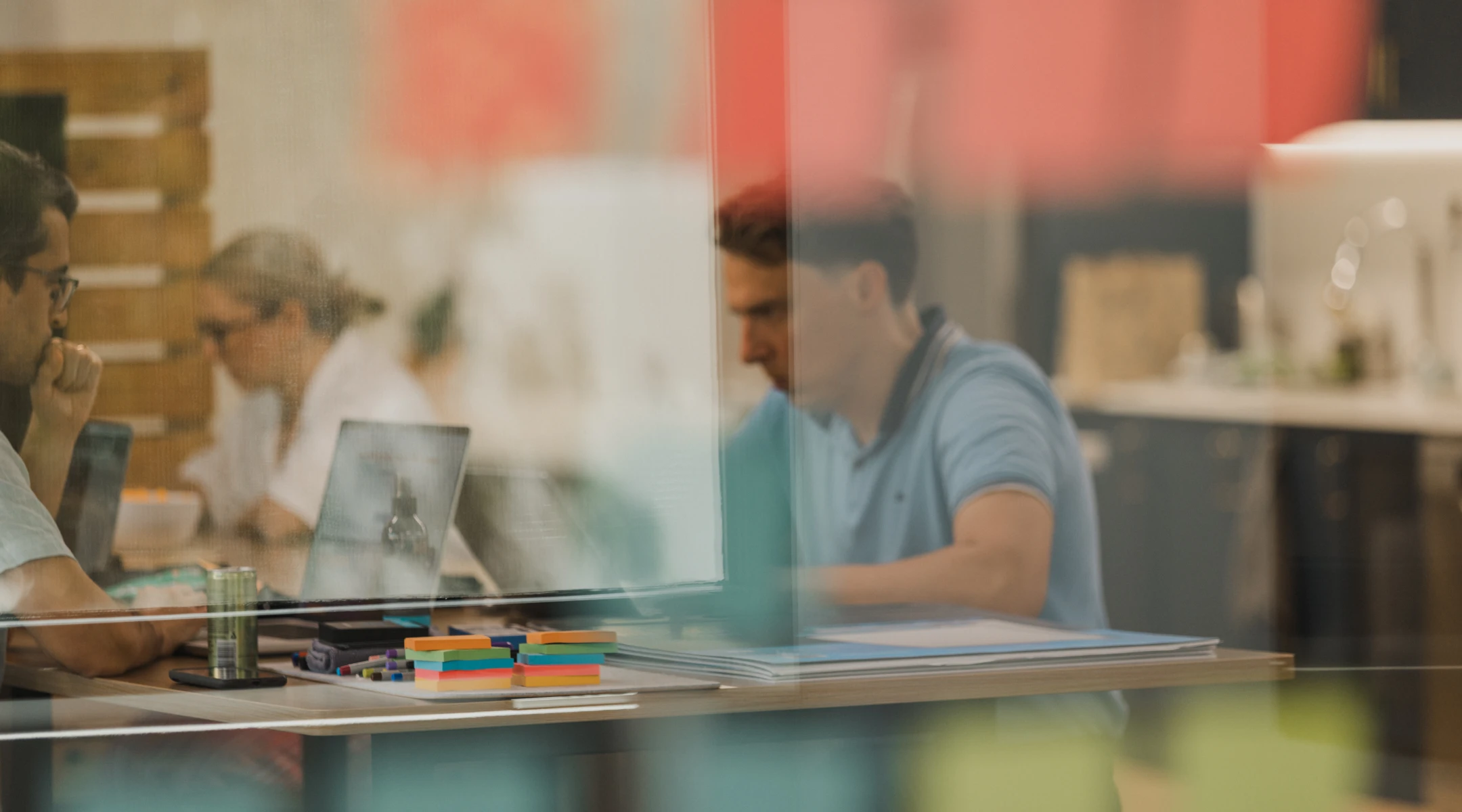 People working in office setting, blurred by glass. Laptops, desk supplies visible. Office workers. Teamwork, collaboration, business.