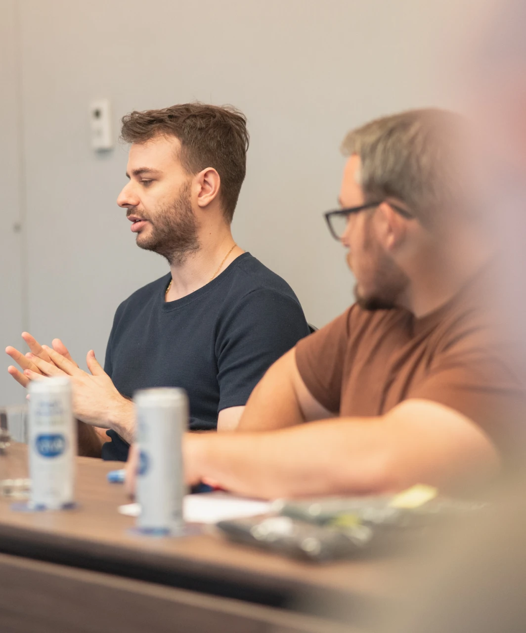 Two men in business meeting, one speaking with hands raised. Cans and papers on table. Collaboration, discussion.