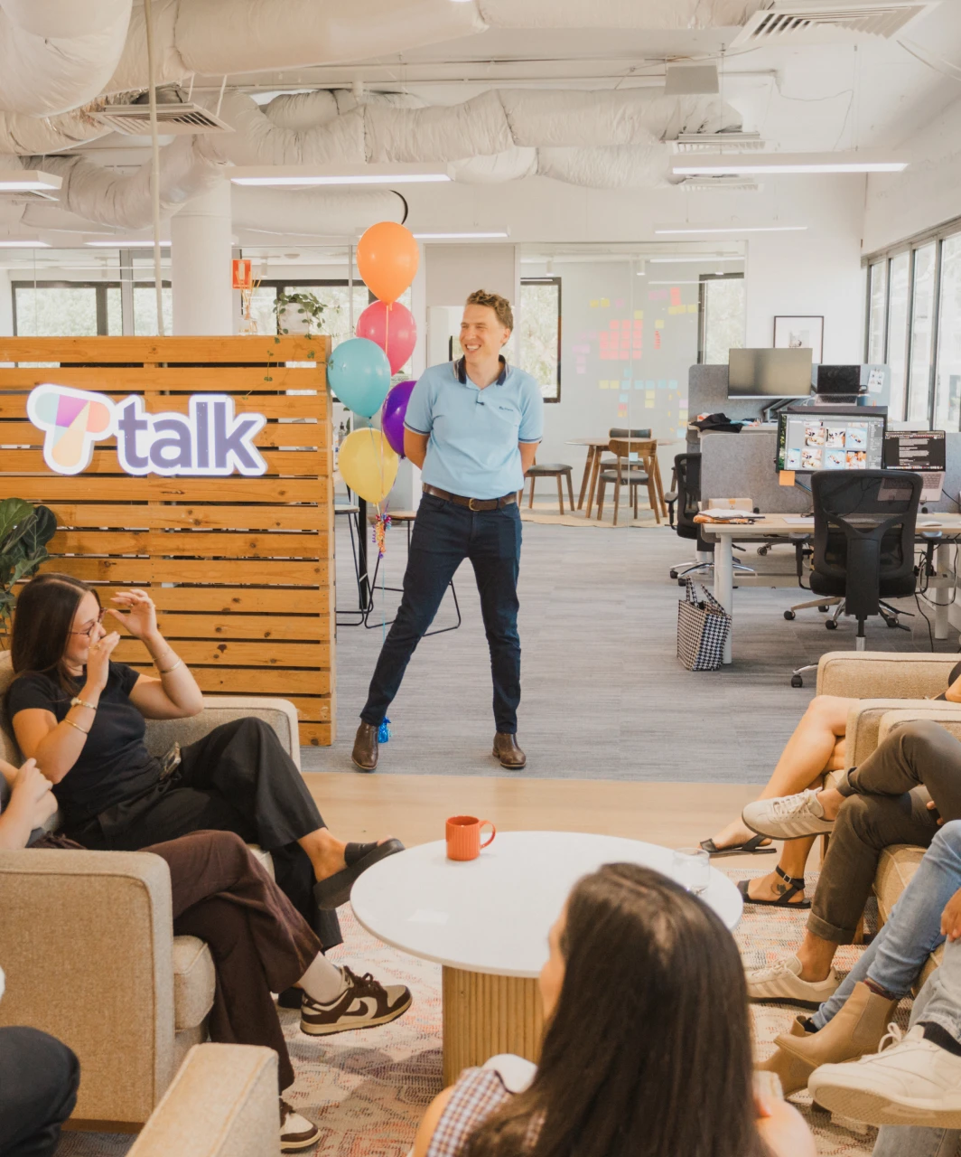 Talk office presentation. Man speaking to colleagues, balloons and Talk logo on wooden wall backdrop. Modern office interior.