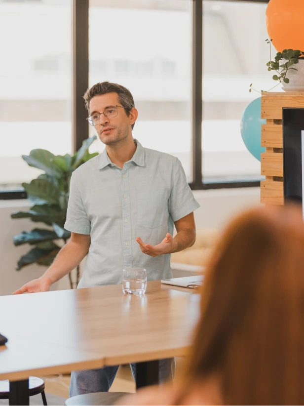 Man giving a presentation at a business meeting, light blue shirt, glasses, gesturing with hands. Corporate event setting.