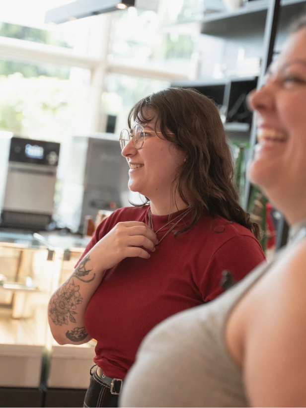 Woman wearing glasses and a red top, smiling. Visible arm tattoos. A second person in background. Natural light.